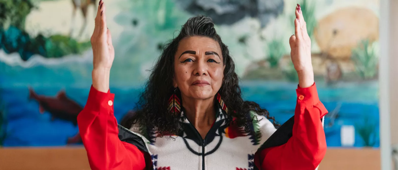 A woman stands with arms raised, wearing a vibrant red shirt and patterned vest. A mural appears in the background