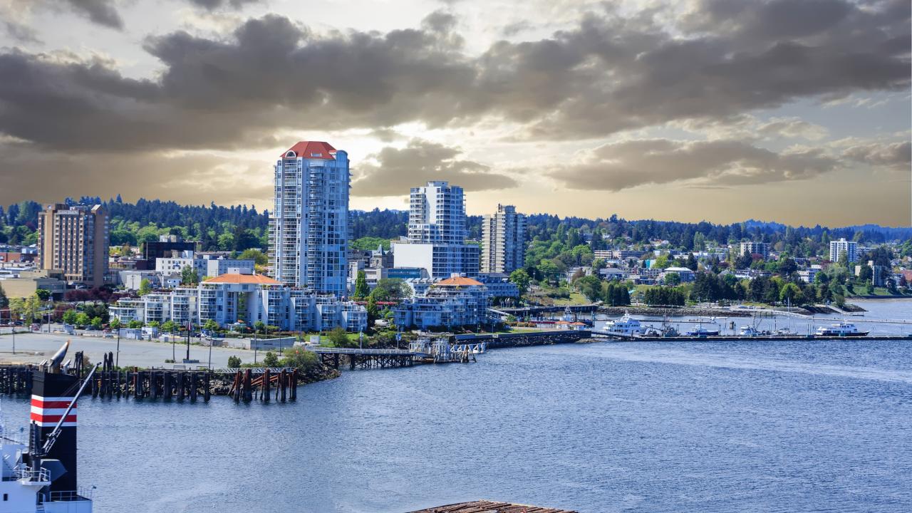Coastal skyline with modern high-rise buildings under a partly cloudy sky. The foreground shows a body of water with a docked ship