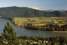 View of a river winding through green fields, bordered by a dense forest and distant snow-capped mountains