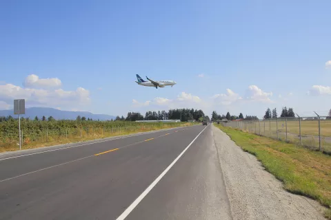 A commercial airplane flies low over a road, surrounded by fields and trees under a clear blue sky with scattered clouds