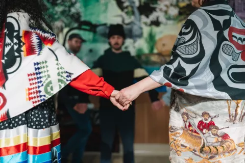 Two people hold hands while wearing brightly colored, patterned shawls. The background shows two people blurred against a mural