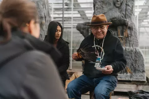 An elderly man in a hat and hoodie holds feathers and a dish, conducting a ceremonial ritual. Two people watch attentively