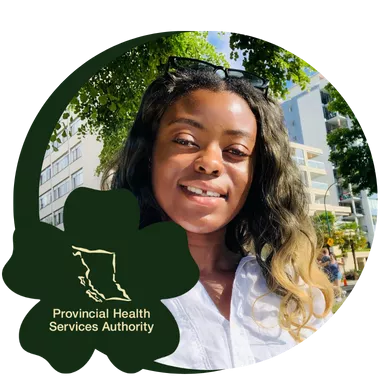 Smiling woman with long hair, wearing a white top and sunglasses on her head, sits on a bench in a sunny park with buildings and trees in the background