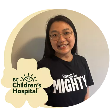 A woman smiles warmly, wearing glasses and a black shirt with the words "Small is Mighty"