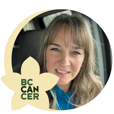 A woman with long brown hair, smiling softly, is seated in a car. She wears a blue shirt and black jacket