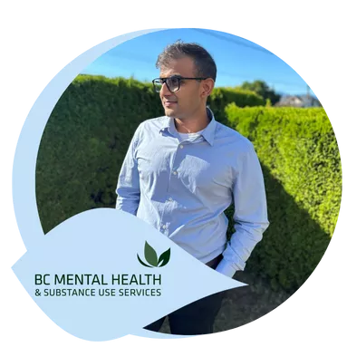 A man wearing glasses and a light blue shirt stands with hands in pockets, smiling in a sunny garden