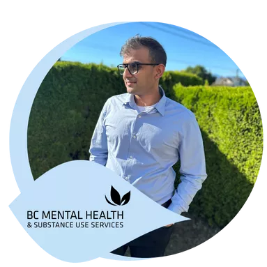 A man wearing glasses and a light blue shirt stands with hands in pockets, smiling in a sunny garden