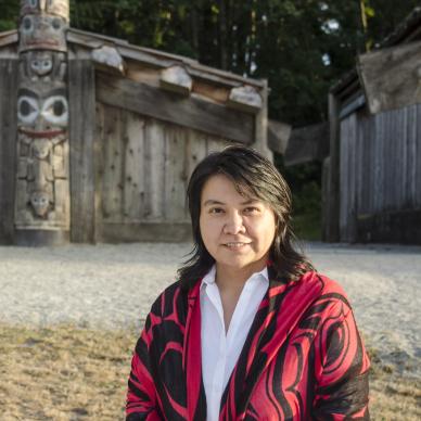 An Indigenous Employed Student Nurse standing in front of a long house and totem pole